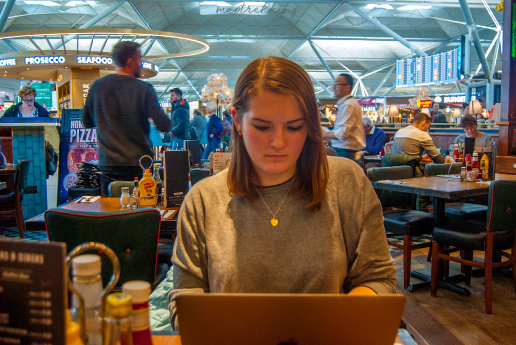 Maria sitting in London Heathrow airport on her laptop. Attempting to figure out our Workaway costs for the first few months. People quickly moving around her in the busy airport.
